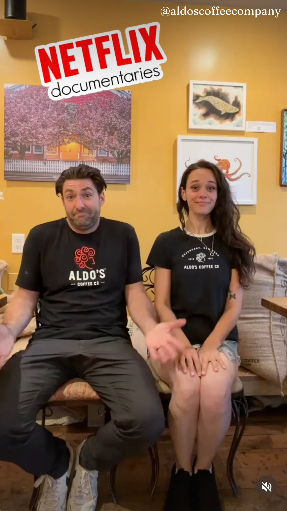 A video still from and Instagram Reel from "@aldoscoffeecompany" showing a man and woman wearing matching "Aldo's Coffee Co." t-shirts sitting on a bench inside a warmly lit coffee shop decorated with framed art and a floral mural, with a red and white "Netflix documentaries" logo overlay at the top.